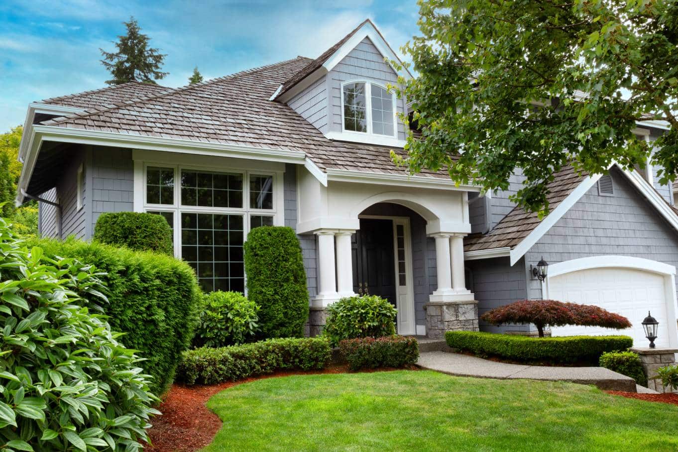 A house with a light blue exterior, a gray cedar roof, and hedges of varying heights in the front yard's landscaping.