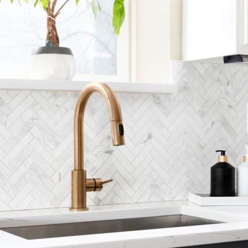 home improvement contractors A golden kitchen sink faucet sits on a gorgeous white countertop. The white kitchen backsplash has a herringbone pattern.
