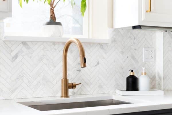 A golden kitchen sink faucet sits on a gorgeous white countertop. The white kitchen backsplash has a herringbone pattern.