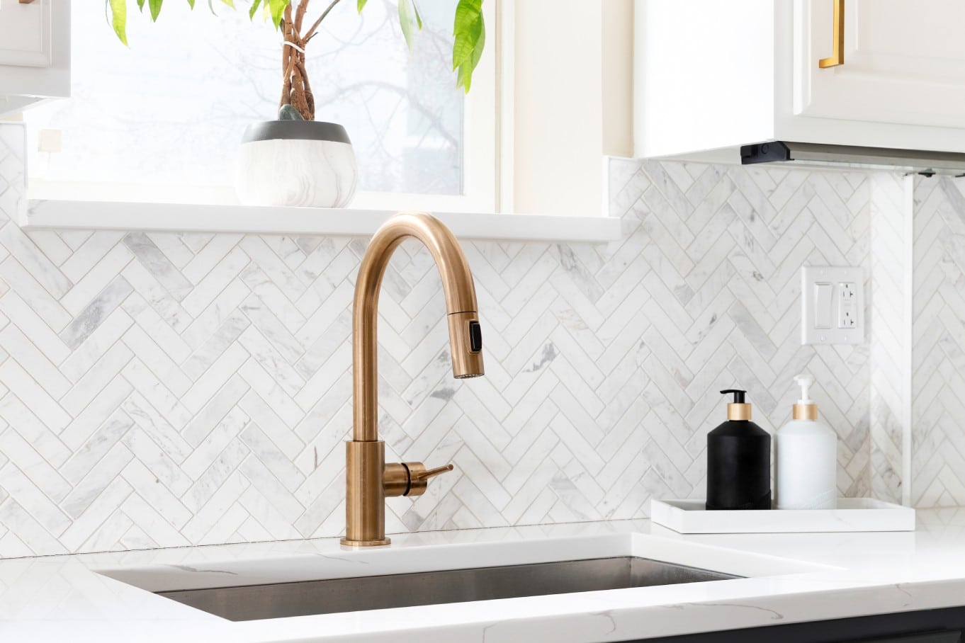 A golden kitchen sink faucet sits on a gorgeous white countertop. The white kitchen backsplash has a herringbone pattern.