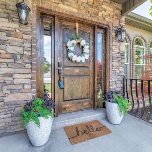 home improvement contractors A stone home with a brown wood front door with a floral wreat, lock box, and welcome mat that says “hello.”