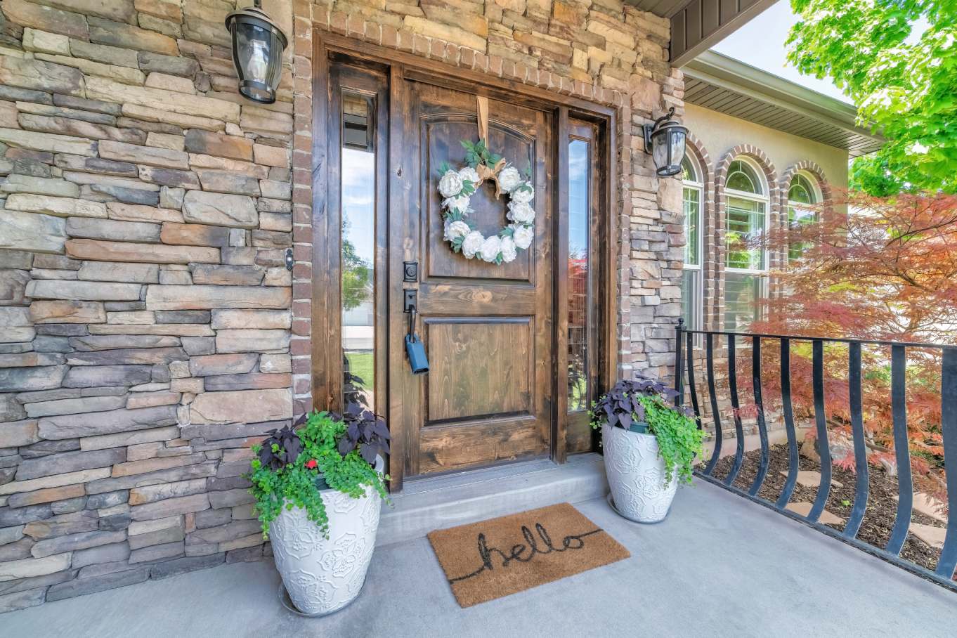 A stone home with a brown wood front door with a floral wreat, lock box, and welcome mat that says “hello.”