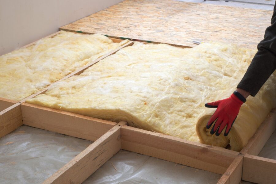 A person wearing red and black gloves and a black jumpsuit is applying insulation to a home's floor.