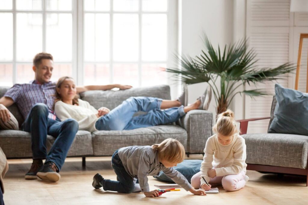 A young girl and boy color together on the floor while their parents sit on the couch watching.