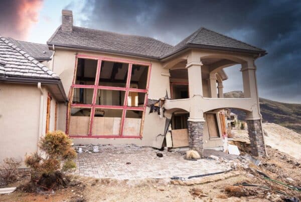 A home with a crumbling foundation that’s suffered significant fire damage. There’s smoke in the sky behind it.