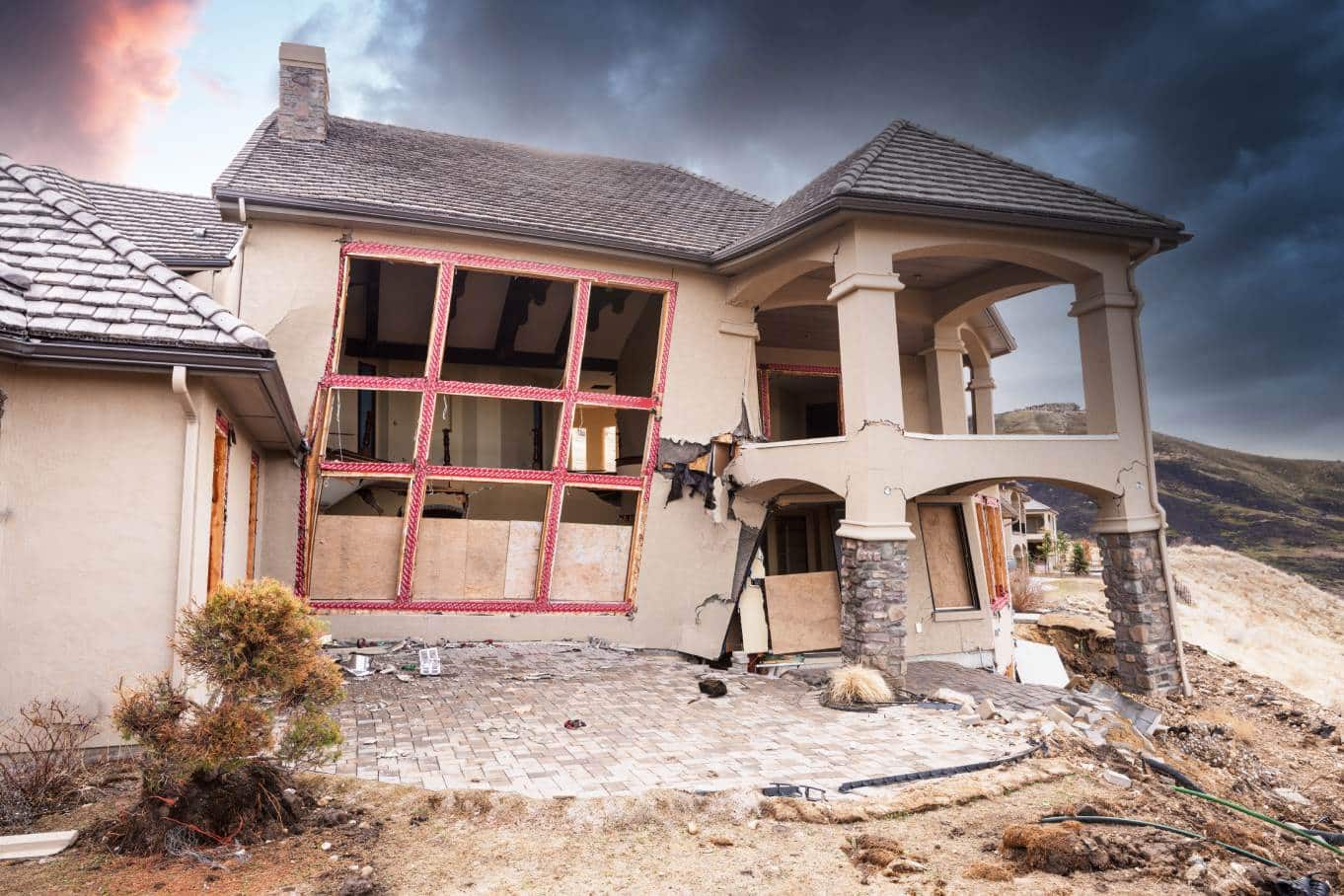 A home with a crumbling foundation that’s suffered significant fire damage. There’s smoke in the sky behind it.