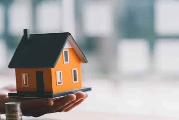 A miniaturized home in the palm of someone's hand is next to three stacks of quarters in varying heights.