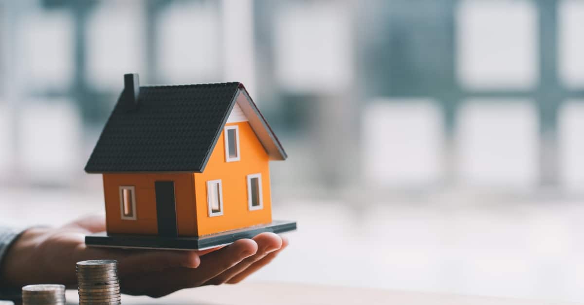 A miniaturized home in the palm of someone's hand is next to three stacks of quarters in varying heights.