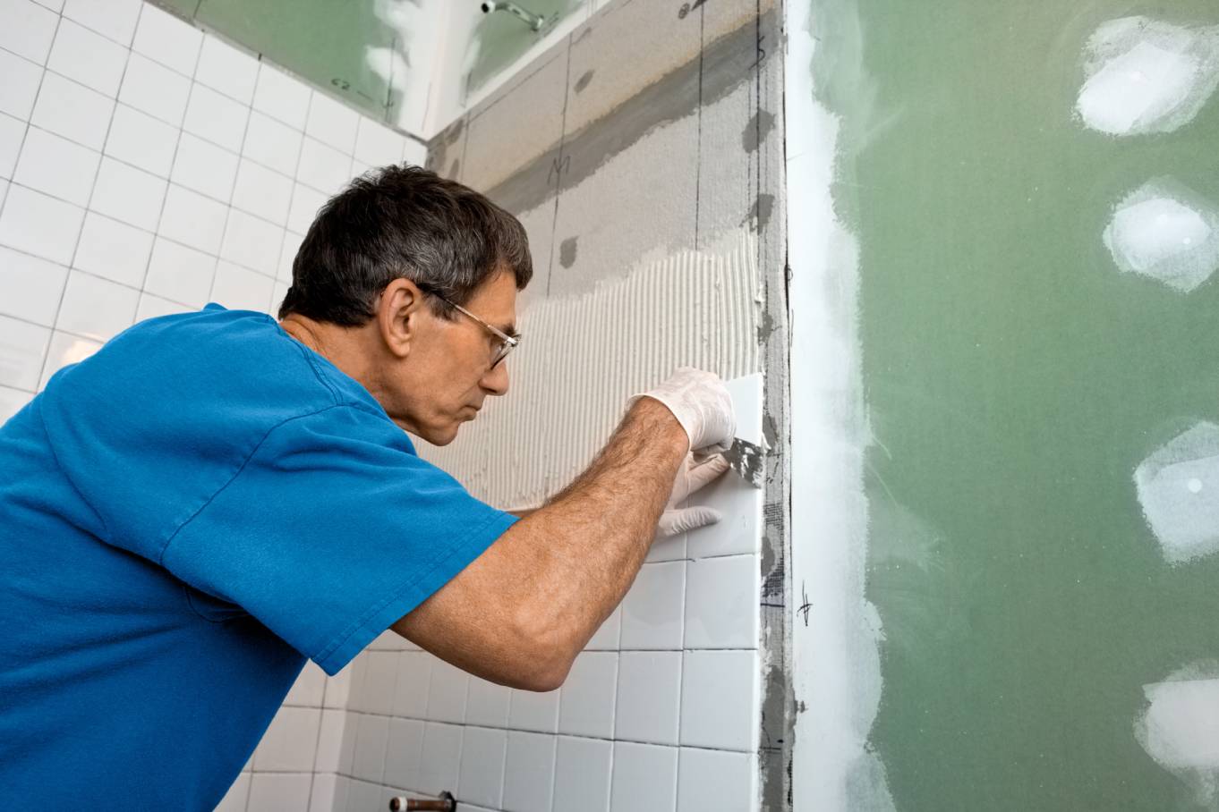 A man in a blue shirt installs white tiles on a bathroom tub enclosure. To the right, there's a green wall with white streaks.