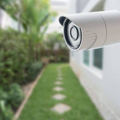 home improvement contractors A close-up shot of a white CCTV security camera on the side of a home. The grassy walkway leading into the back is blurred.