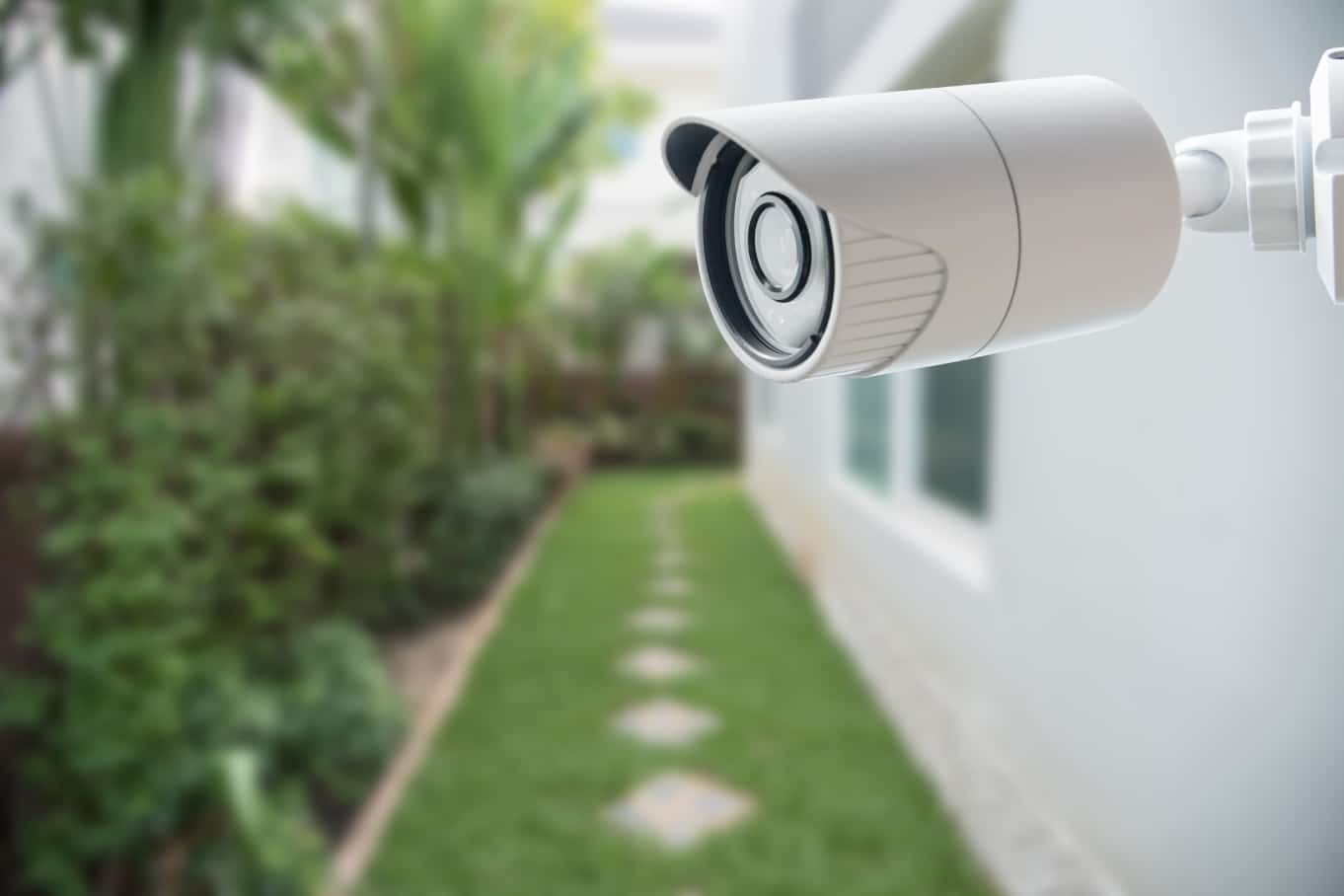 A close-up shot of a white CCTV security camera on the side of a home. The grassy walkway leading into the back is blurred.