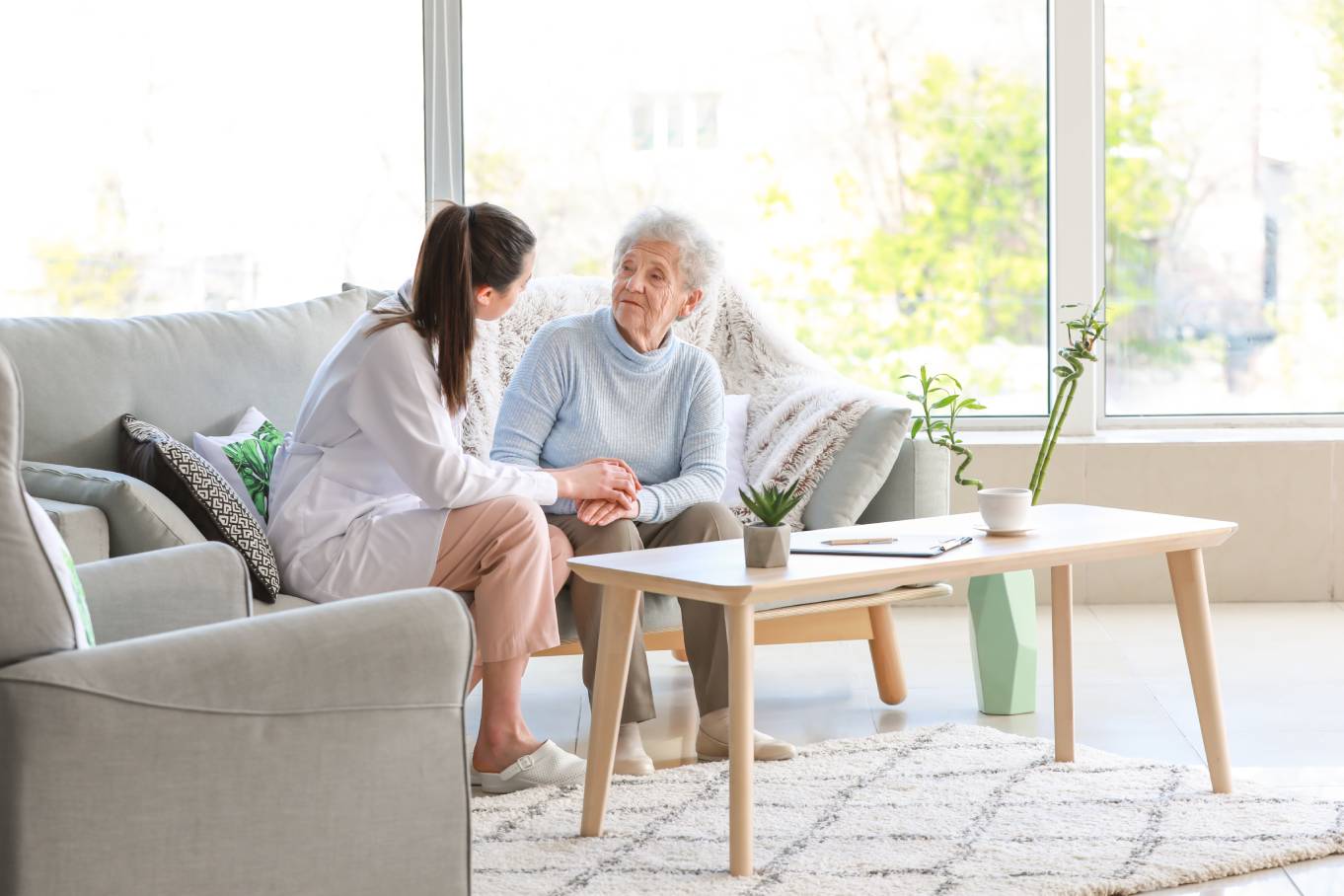 A senior loved one sitting with a family member having a conversation on a gray sofa. There's a coffee table in front.