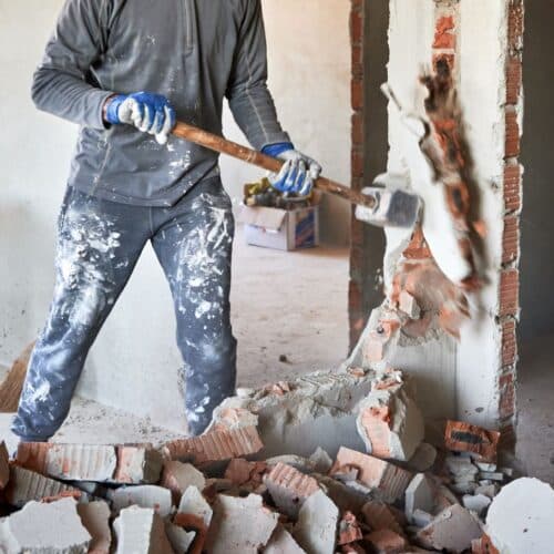 home improvement contractors A man wearing a dusty gray shirt and jeans and holding a sledgehammer standing in rubble and knocking a wall down.