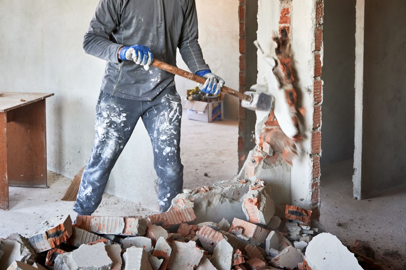 A man wearing a dusty gray shirt and jeans and holding a sledgehammer standing in rubble and knocking a wall down.