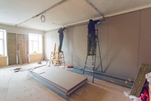 Two people stand on ladders as they work on the plasterboard of a tall wall inside a room with numerous construction materials.