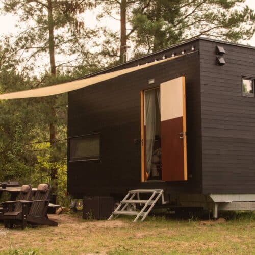 home improvement contractors A black tiny home built on top of a trailer bed sitting outside in the woods next to a fire pit with a canopy over it.