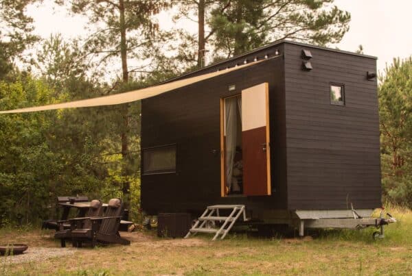 A black tiny home built on top of a trailer bed sitting outside in the woods next to a fire pit with a canopy over it.
