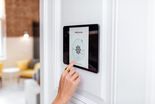 A right hand with a gold ring on the index finger is touching a tablet's screen hanging on a home wall.