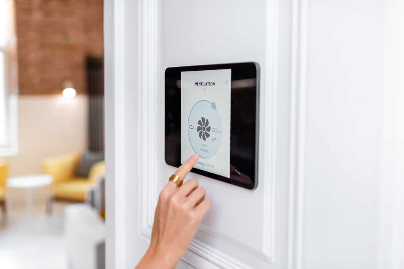 A right hand with a gold ring on the index finger is touching a tablet's screen hanging on a home wall.