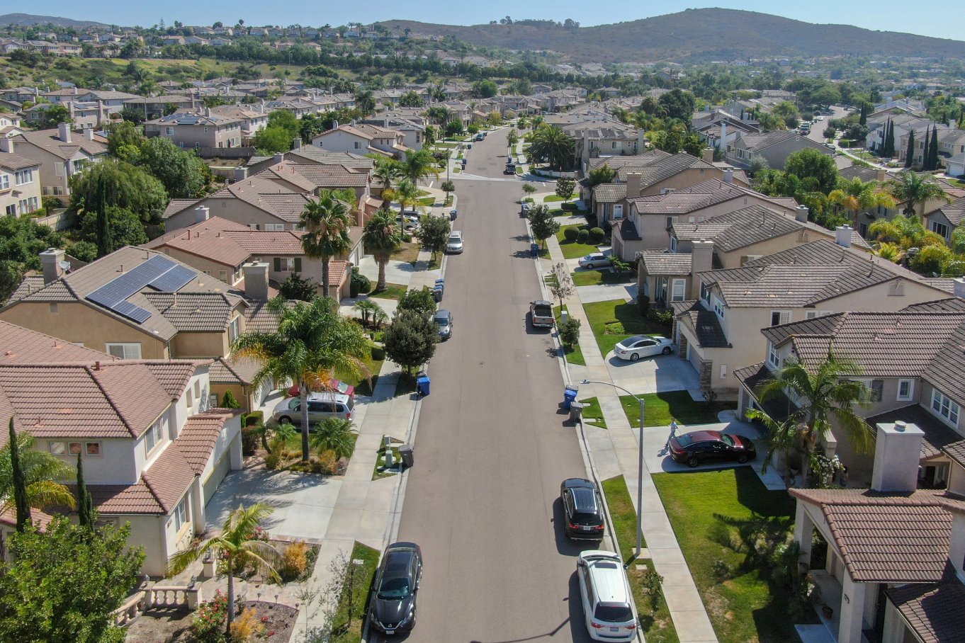A suburban neighborhood is filled with houses. There are several cars parked in front of the homes on a sunny day.