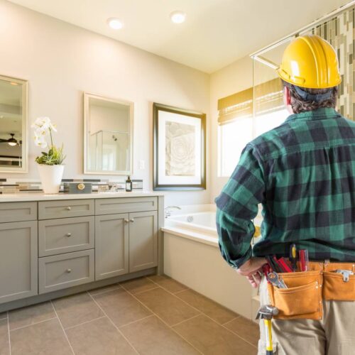 home improvement contractors A man wearing a yellow hard hat and a plaid shirt standing in a large, brightly lit bathroom with white walls.