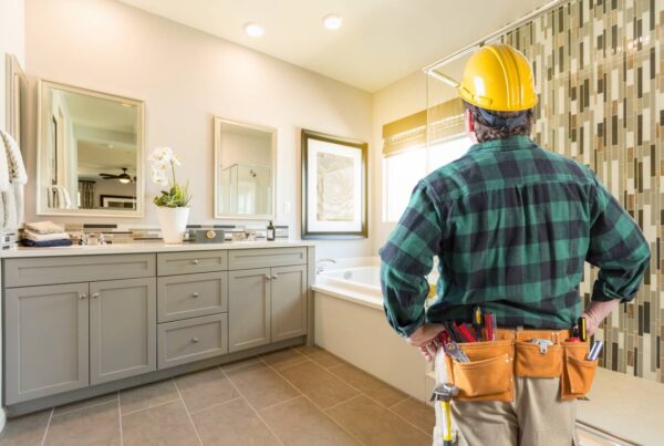 A man wearing a yellow hard hat and a plaid shirt standing in a large, brightly lit bathroom with white walls.