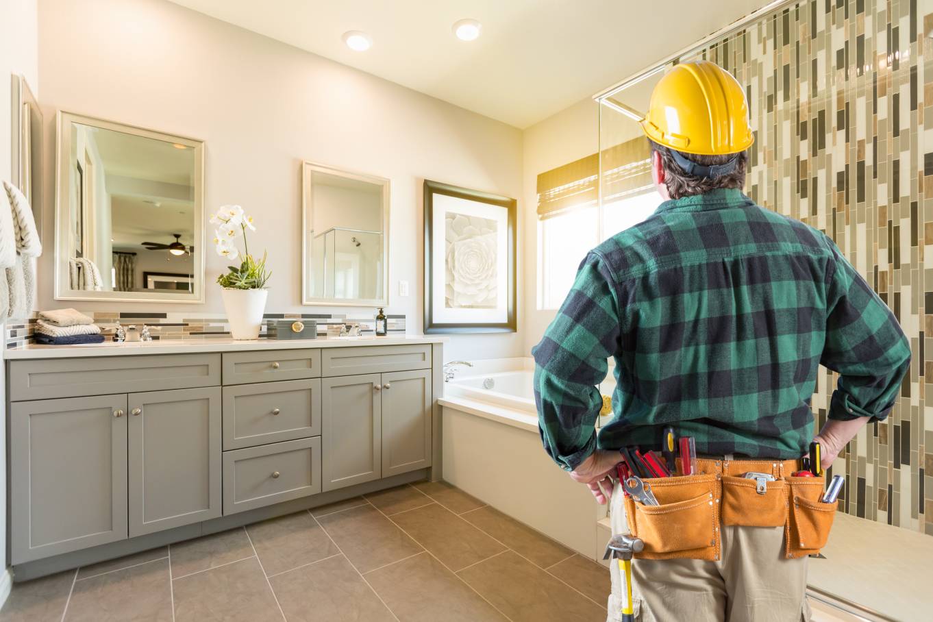 A man wearing a yellow hard hat and a plaid shirt standing in a large, brightly lit bathroom with white walls.