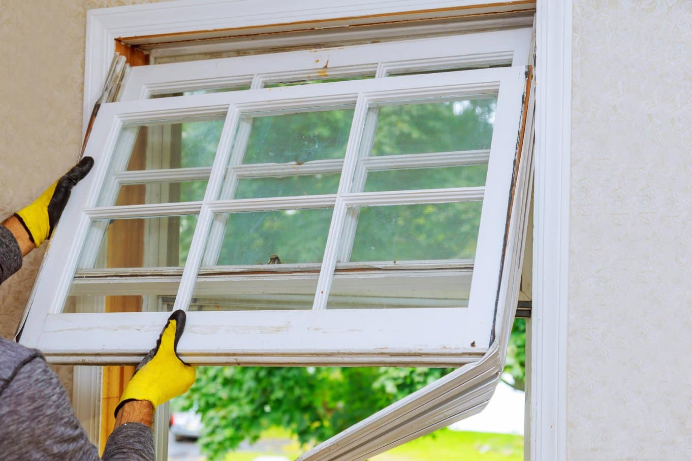 A man wearing yellow gloves is removing an old window with a white frame that appears to need replacing.