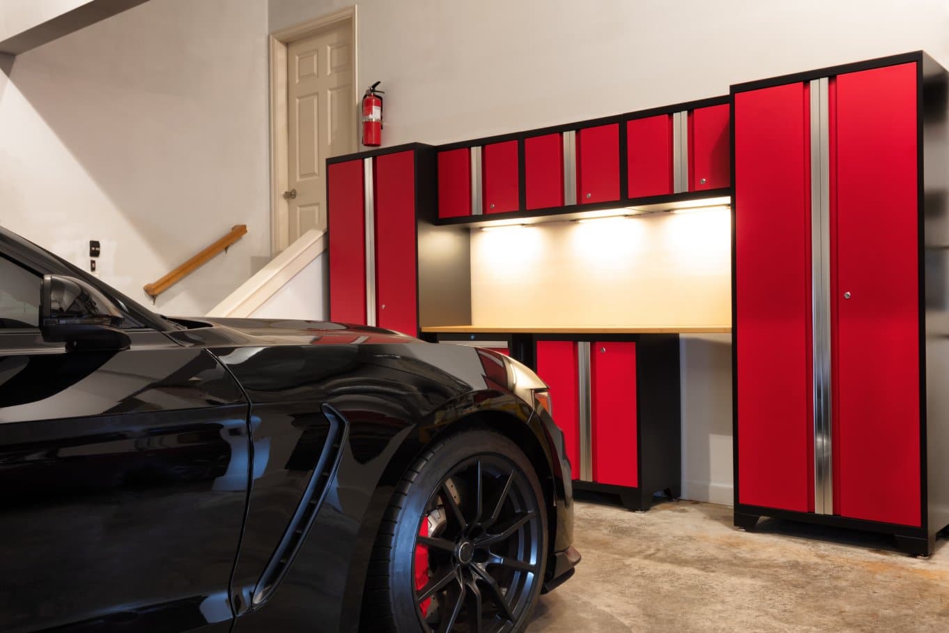 A nice, clean home garage with a black car parked close to the entry door. There is a red cabinet unit in front of the car.