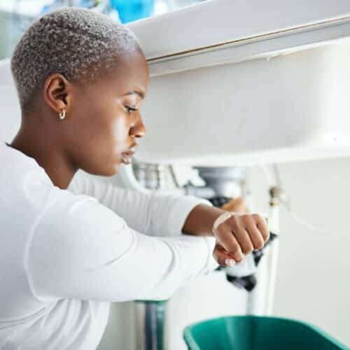 home improvement contractors A woman wearing a white shirt kneels under a bathroom sink with a green bucket underneath it as she looks at her watch.