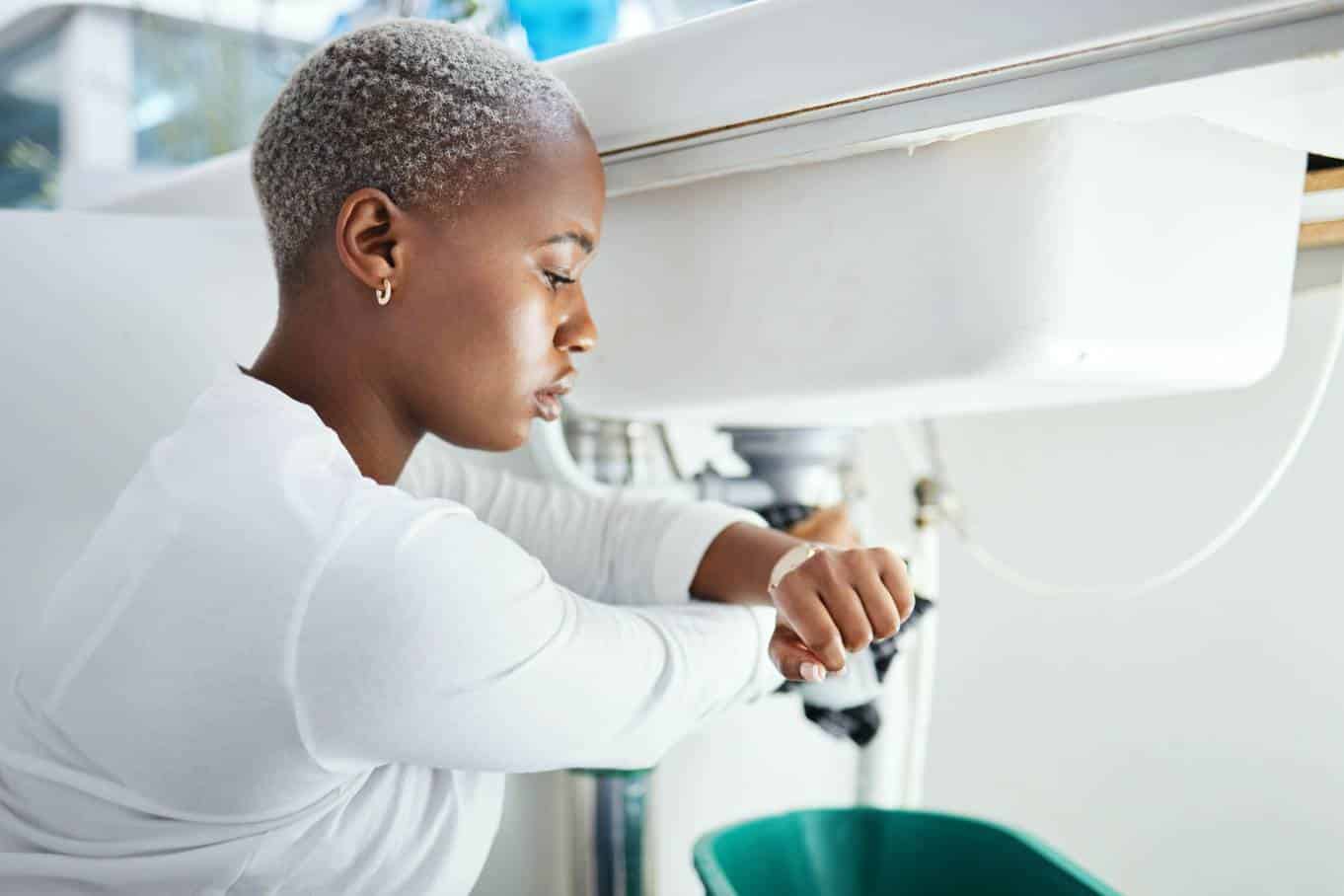 A woman wearing a white shirt kneels under a bathroom sink with a green bucket underneath it as she looks at her watch.