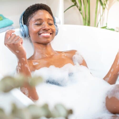 home improvement contractors A smiling Black woman covered in bubbles in a bathtub. She is wearing headphones and surrounded by plants.