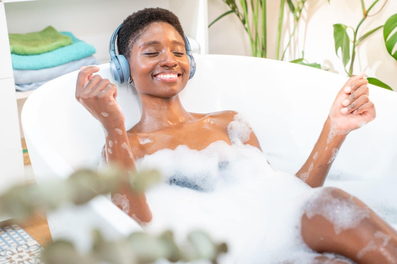 A smiling Black woman covered in bubbles in a bathtub. She is wearing headphones and surrounded by plants.
