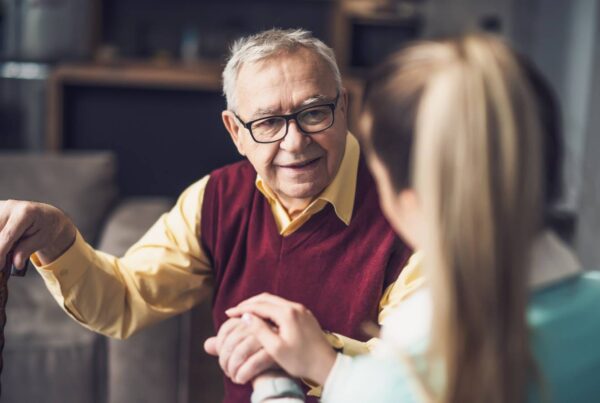An older man wearing glasses and holding onto a cane in his left hand looks ahead at a woman who holds his hand.