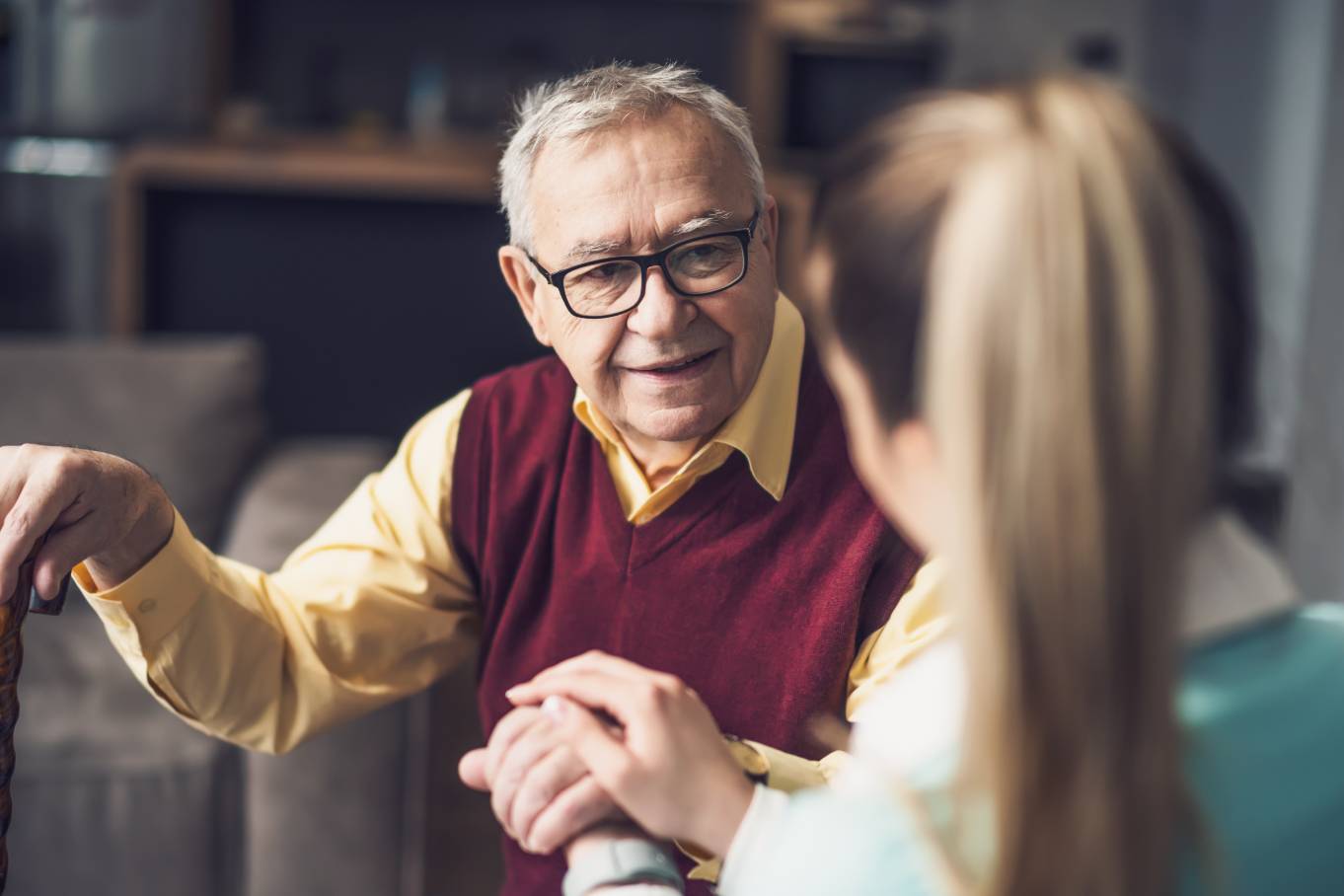 An older man wearing glasses and holding onto a cane in his left hand looks ahead at a woman who holds his hand.