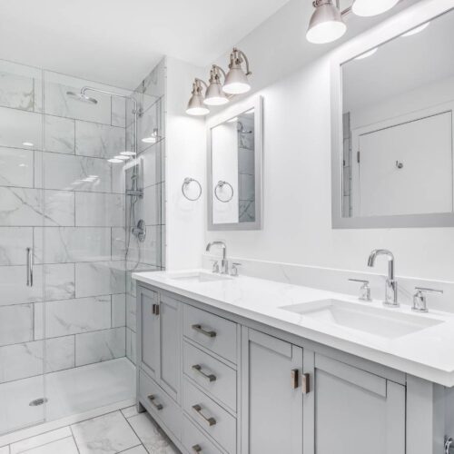 home improvement contractors A modern bathroom contains a gray vanity with double sinks, a walk-in shower with a glass door, and silver fixtures.