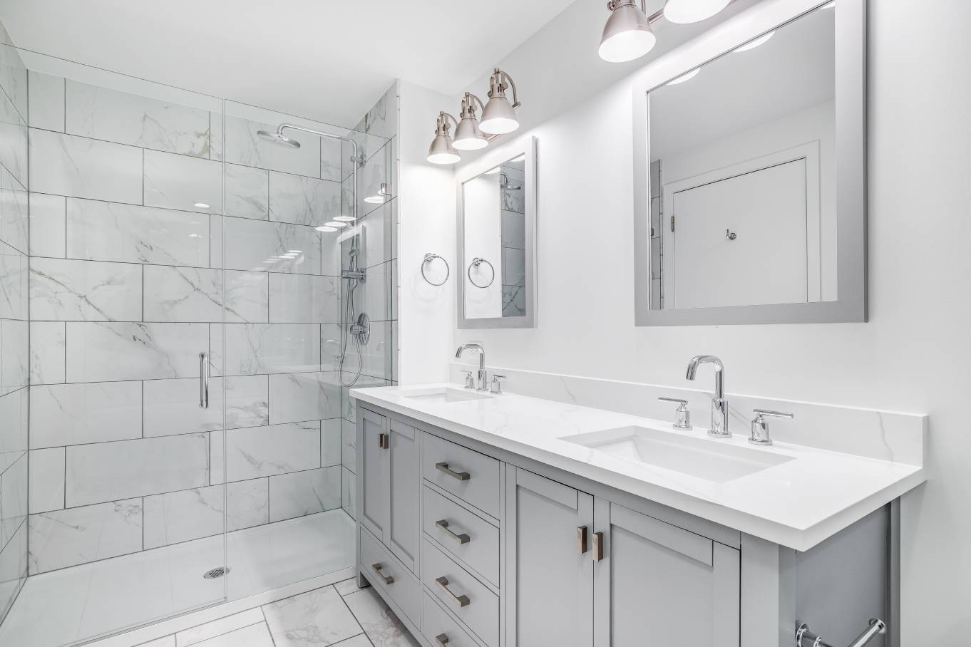 A modern bathroom contains a gray vanity with double sinks, a walk-in shower with a glass door, and silver fixtures.