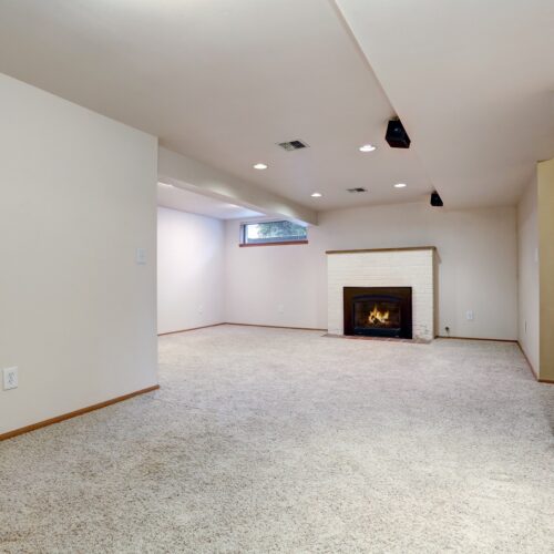 home improvement contractors An empty, spacious room with white walls, brown trim, and grey carpeting with a black fireplace on the other side of the room.