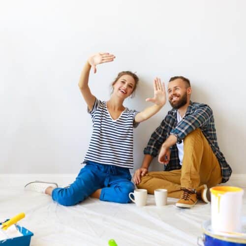 home improvement contractors A smiling man and woman sit on a tarp-covered floor surrounded by painting supplies, a ladder, and two coffee mugs.
