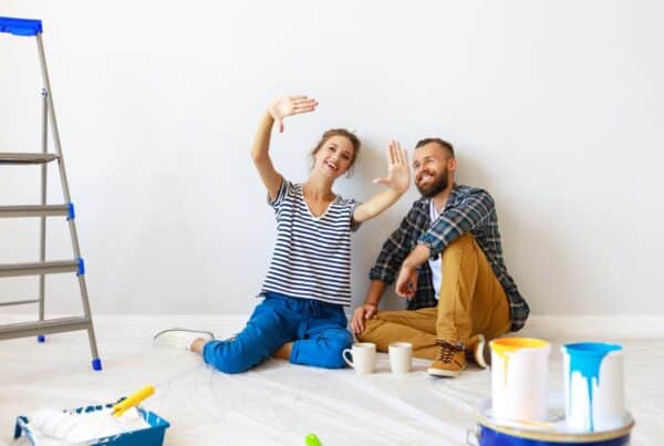 A smiling man and woman sit on a tarp-covered floor surrounded by painting supplies, a ladder, and two coffee mugs.
