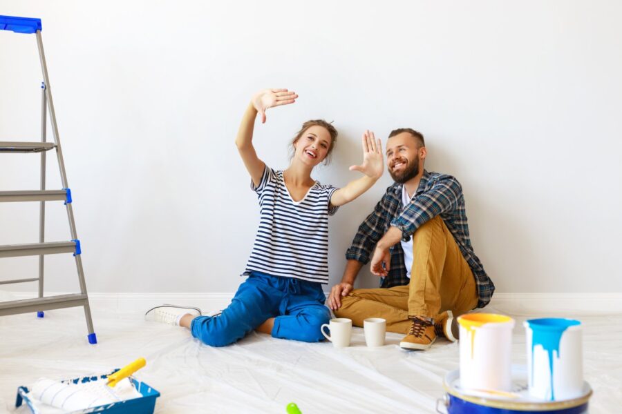 A smiling man and woman sit on a tarp-covered floor surrounded by painting supplies, a ladder, and two coffee mugs.