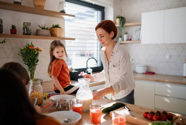 A mother and her three children are preparing breakfast in the kitchen at home. The youngest is sitting on the counter.