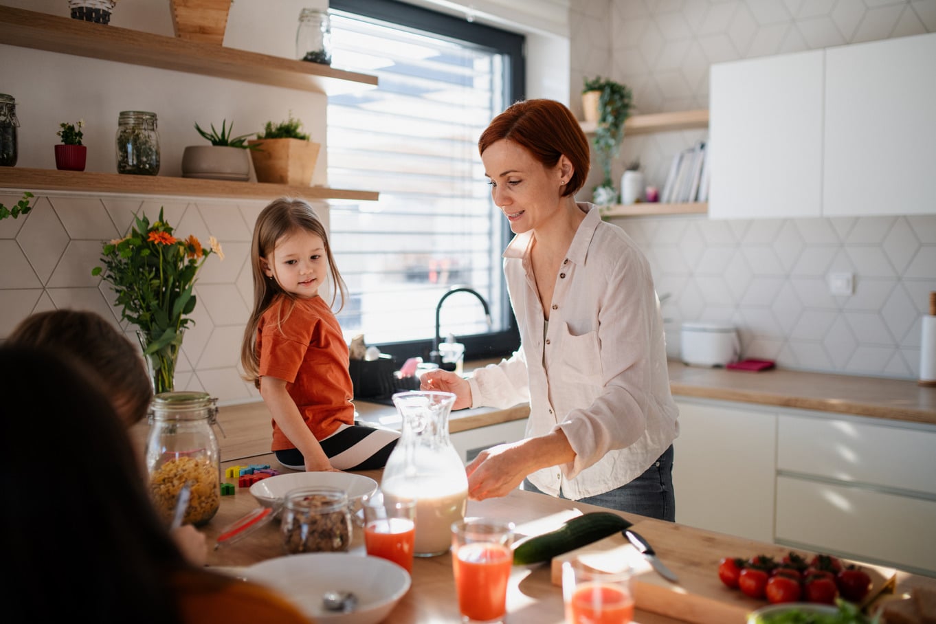 A mother and her three children are preparing breakfast in the kitchen at home. The youngest is sitting on the counter.