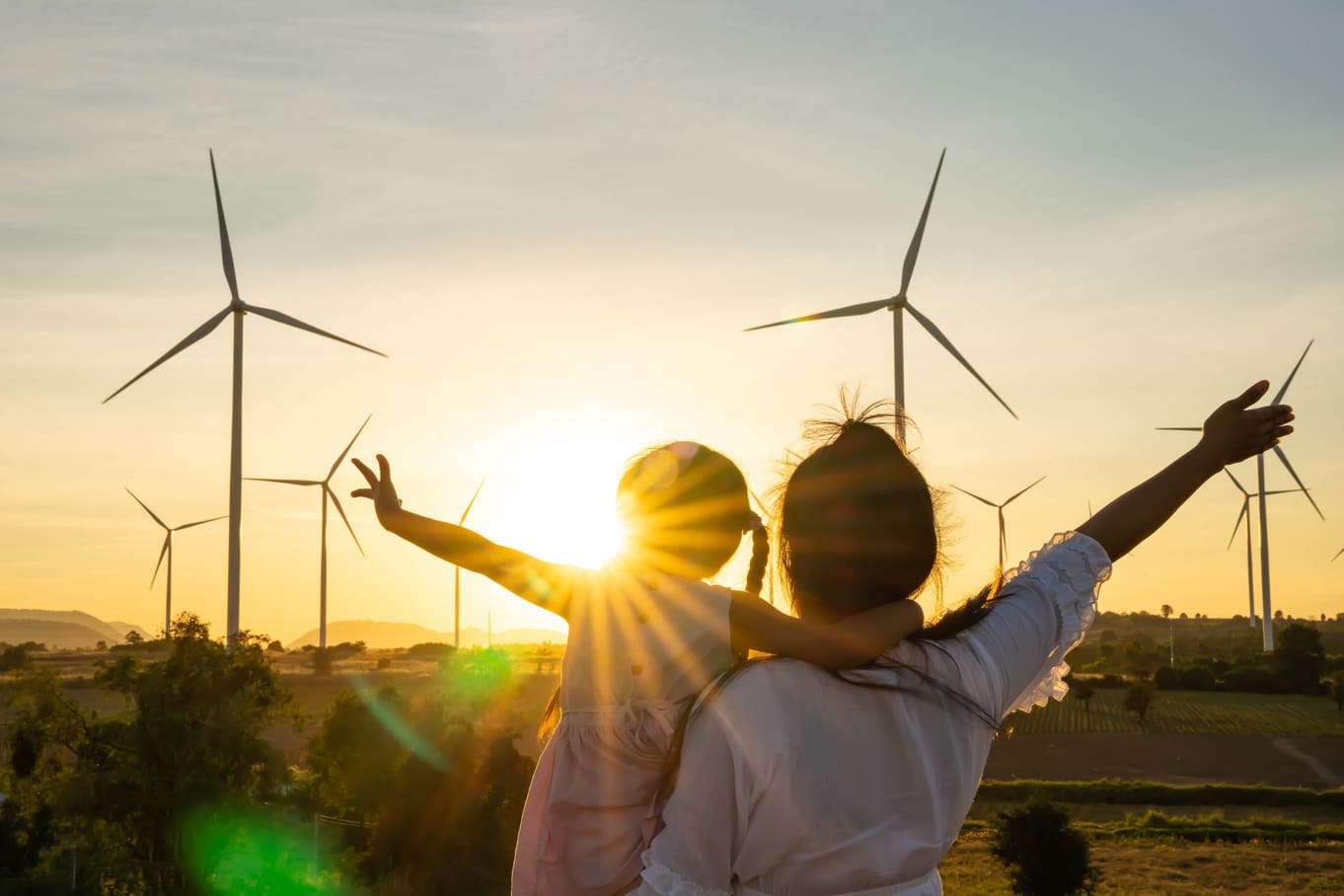 A woman holding up a little girl with her arms out toward the sun in front of a field of wind turbines.