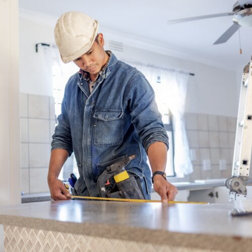 helloproject A handyman wearing a white hard hat measures a kitchen counter next to a ladder under a grey ceiling fan.