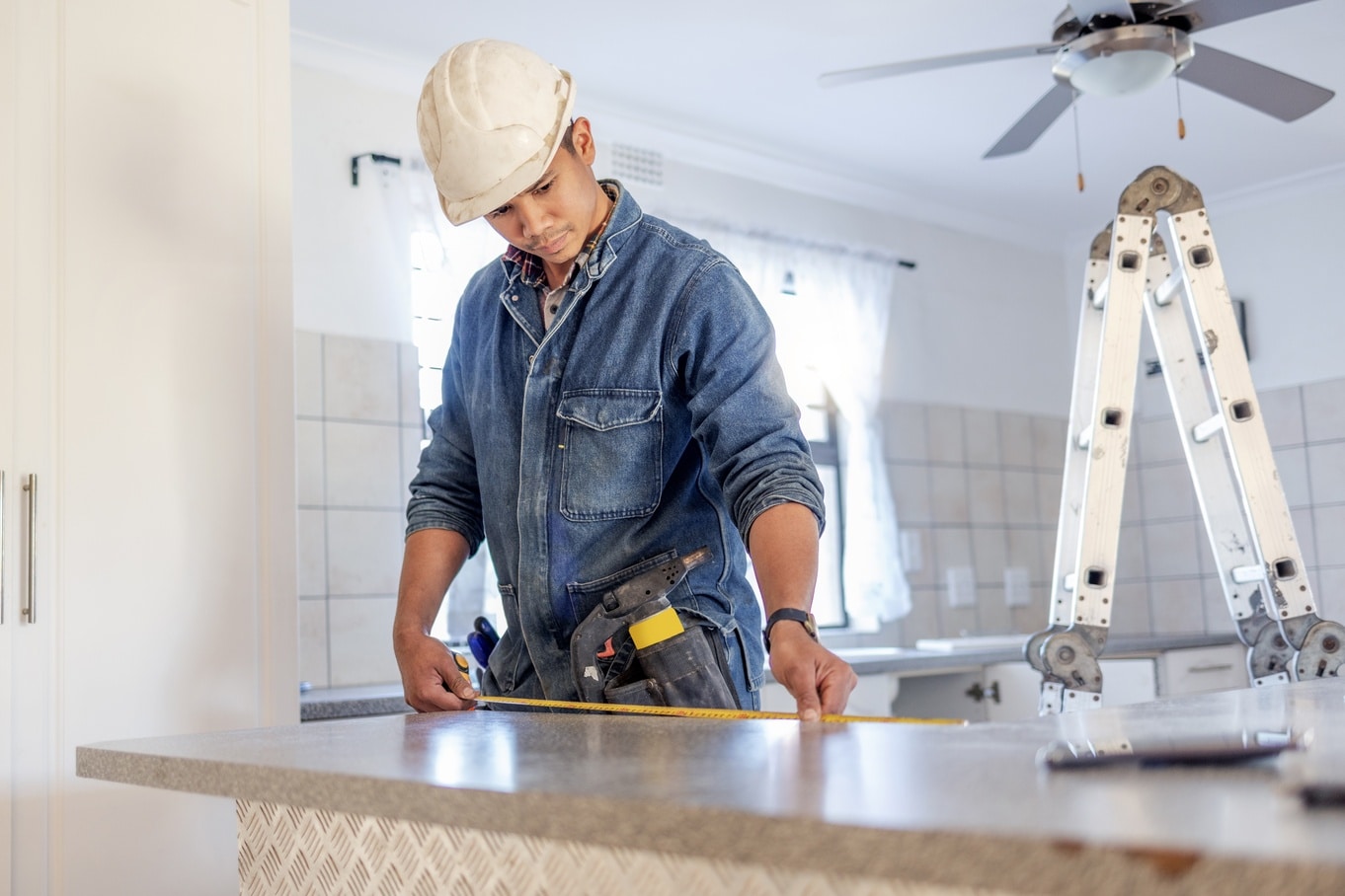 A handyman wearing a white hard hat measures a kitchen counter next to a ladder under a grey ceiling fan.