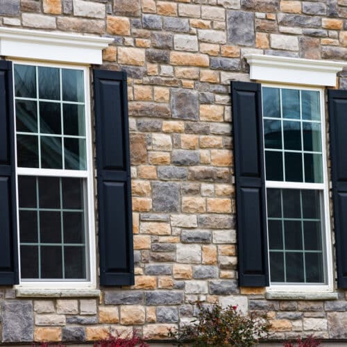 home improvement contractors A close-up view shows a gray and tan stone home with two tall windows. Black shutters sit on either side of the windows.