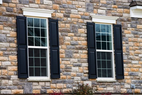 A close-up view shows a gray and tan stone home with two tall windows. Black shutters sit on either side of the windows.