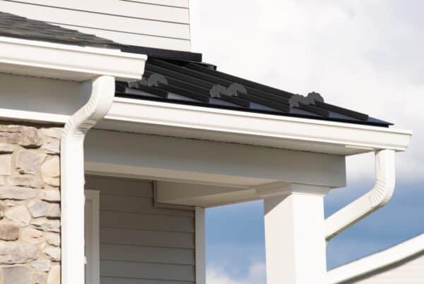 The corner of a home's roof has white gutters, a white downspout, black metal roofing, and gray shingles.