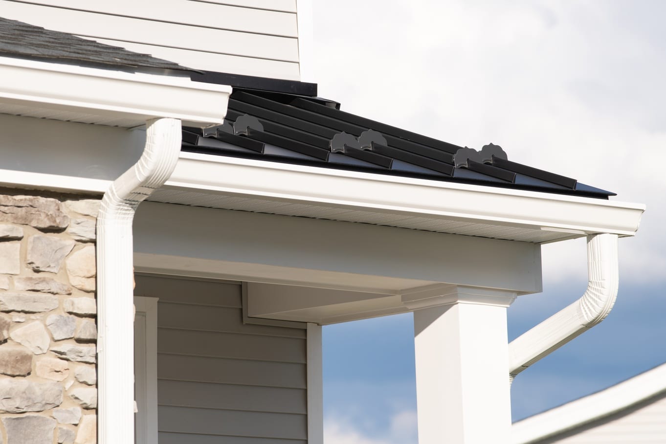 The corner of a home's roof has white gutters, a white downspout, black metal roofing, and gray shingles.